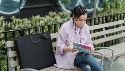 Woman artist sketching on a park bench with a black portfolio beside her.
