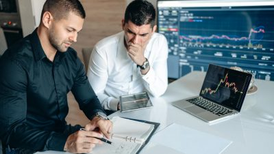 Two businessmen reviewing financial charts on a laptop and tablet in a modern office setting.