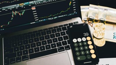 Laptop screen with trading charts, calculator app, Euros and Bitcoin coins on desk.