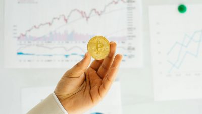 A hand holding a Bitcoin coin against financial charts depicting market trends.