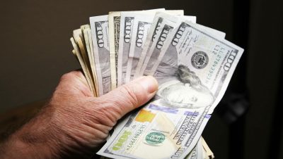 Close-up of a hand holding multiple hundred dollar bills on a dark background.