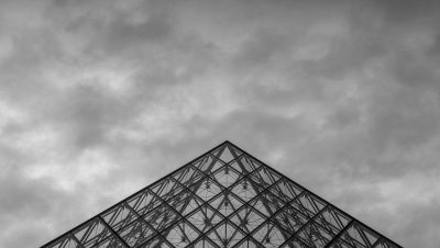 Dramatic black and white shot of the Louvre Pyramid in Paris with cloudy skies.