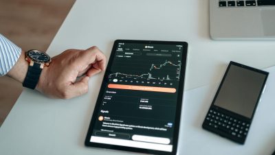 Close-up of a digital tablet displaying a bitcoin chart, with a hand nearby on a white table.