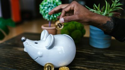 Close-up of hand placing bitcoin into a mouse-shaped piggy bank on a table.